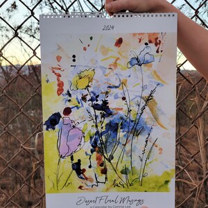 Two hands holding up a 2024 Desert Floral Musings Ledger style wall calendar in front of a chain link fence with a sunset in the background. The Image on the front of the calendar is an abstract floral painting with blue, yellow, and purple flowers.
