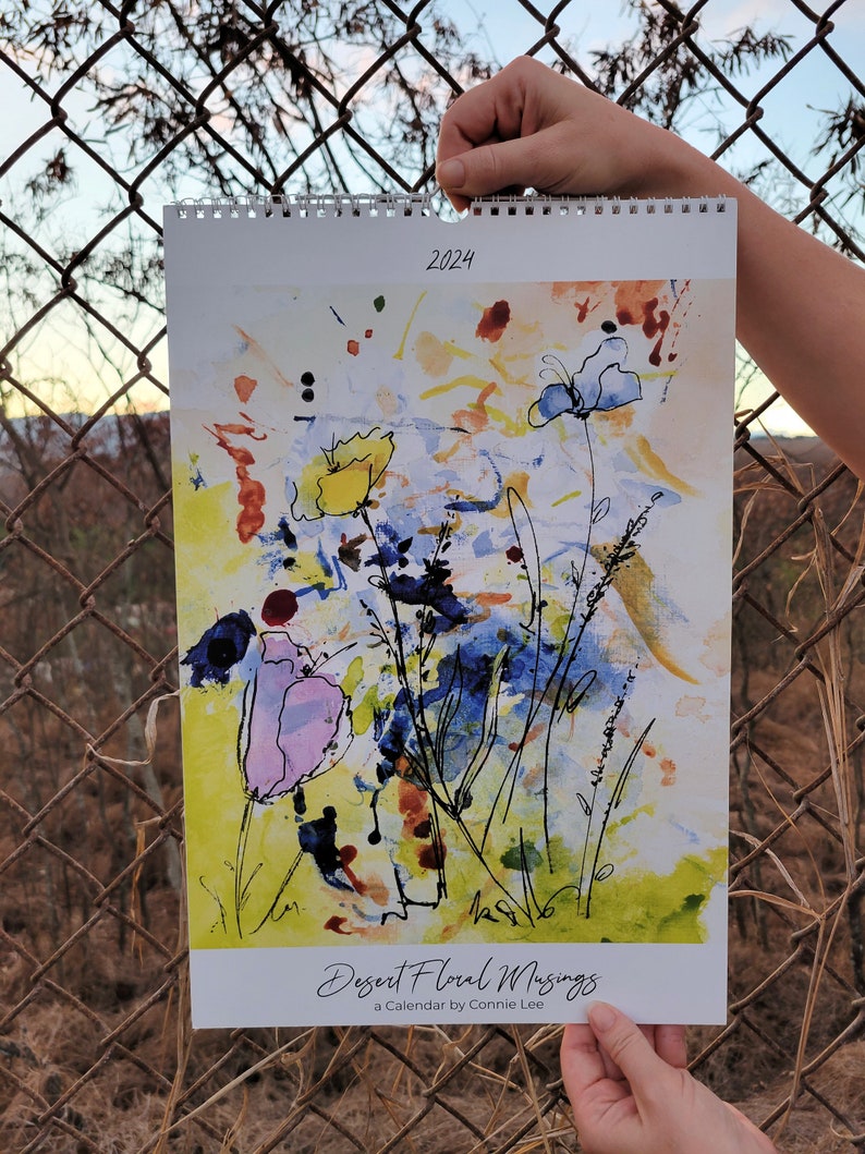 Two hands holding up a 2024 Desert Floral Musings Ledger style wall calendar in front of a chain link fence with a sunset in the background. The Image on the front of the calendar is an abstract floral painting with blue, yellow, and purple flowers.