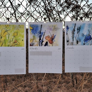 Three 2024 wall calendars hanging on a chain link fence showcasing the abstract floral images for the months of October, November, and December. They are yellow, blue, and orange.