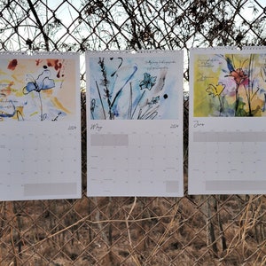 Three 2024 wall calendars hanging on a chain link fence showcasing the abstract floral images for the months of April, May, and June. They are primarily blue and orange/yellow.