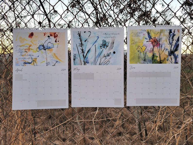 Three 2024 wall calendars hanging on a chain link fence showcasing the abstract floral images for the months of April, May, and June. They are primarily blue and orange/yellow.