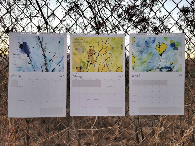 Three 2024 wall calendars hanging on a chain link fence showcasing the abstract floral images for the months of January, February, and March. They are primarily blue and yellow.