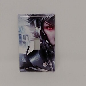May include: A light switch cover featuring a close-up of a character with a silver and black helmet and a red eye. The background is a blurry city scene.