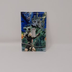May include: A light switch cover featuring a colorful illustration of a man with a whip fighting a vampire in front of a castle. The image is from the Castlevania video game.