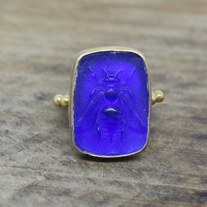 Pode incluir: Um anel de ouro com uma configuração de vidro azul com um design de abelha gravada.