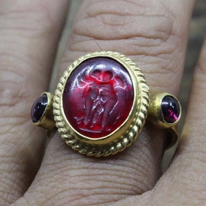 Può includere: Un anello d'oro con una pietra ovale rossa incastonata in un bordo a forma di corda. Due pietre rosse più piccole sono incastonate su entrambi i lati della pietra principale.