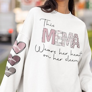 May include: White sweatshirt with the text "This Mema wears her heart on her sleeve" and three pink hearts with names on the sleeve.
