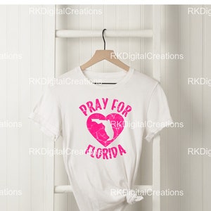 Könnte beinhalten: Weißes T-Shirt mit einem pinkfarbenen Herzen, das die Umrisse des Bundesstaates Florida zeigt. Der Text "Pray for Florida" ist auf das Shirt gedruckt.