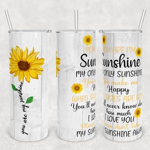 May include: A white tumbler with a wood grain pattern and a sunflower graphic. The tumbler has the text "You are my sunshine" on the side. The text "You are my sunshine" is repeated on the other side of the tumbler, with the text "You make me happy" and "When skies are gray, you'll never know how much I love you. Please don't take my sunshine away."