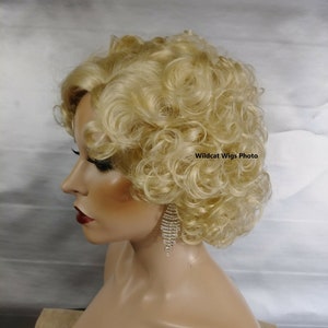 May include: A blonde wig with tight curls styled on a mannequin head. The wig is a light, golden color and has a voluminous, curly texture. The mannequin is wearing a pair of dangling, silver rhinestone earrings. The text "Wildcat Wigs Photo" is visible.