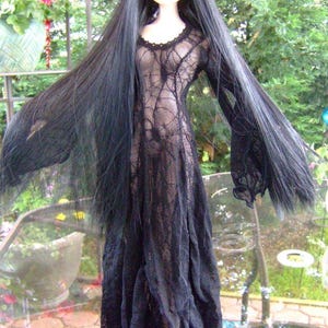 May include: A doll with long black hair and bangs, wearing a black lace dress with long sleeves. The dress has a delicate, web-like pattern. The doll is posed outdoors, with a blurred background of greenery.