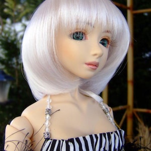 May include: A doll with a white wig and green eyes, wearing a black and white striped dress. The doll's dress has beaded straps and black feather accents. The image is labeled "Wildcat Wigs Photo".