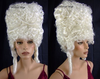 May include: A white, curly wig with a high, rectangular shape, adorned with a strand of pearls. The wig features long, curled sections framing the face. The text "Wildcat Wigs" is visible in the upper left corner.