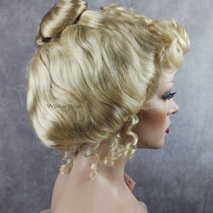 May include: A blonde wig with a bun on top and curls on the sides. The wig is styled in a classic updo. The wig is on a mannequin head. The text "Wildcat Wigs" is visible on the wig.