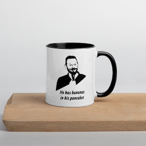Puede incluir: Una taza de cerámica blanca con borde y asa negros. La taza presenta una ilustración en blanco y negro de un hombre con el texto "He has bananas in his pancakes".