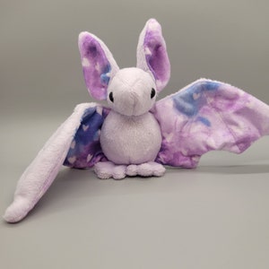 May include: A plush purple bat with a white and blue tie-dye pattern on its wings. The bat has a small, round body and large, floppy ears.