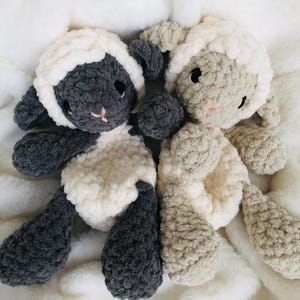 May include: Two soft toy sheep, one grey and white, the other beige and white. Both have black eyes and pink noses. The toys are made of a soft, textured material and are lying on a white, fluffy surface.