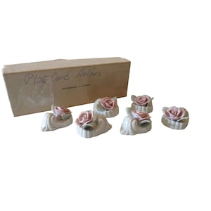 May include: A set of six ceramic place card holders, each featuring a delicate pink rose. The holders are off-white with a scalloped design. The box reads "Place Card Holders" and "Strawbridge & Clothier".