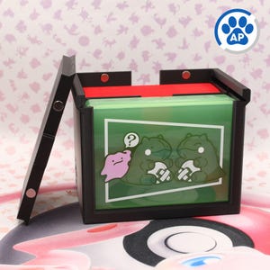 Könnte beinhalten: Schwarze und rote Kartenaufbewahrungsbox mit Klappdeckel. Die Box hat einen grünen Innenraum mit einer Cartoon-Grafik von zwei grünen Dinosauriern und einem rosa Klecks. Ein blaues Pfotenabdruck-Logo ist sichtbar.