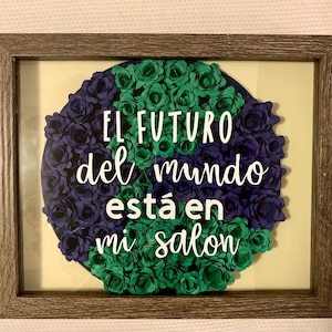 以下が含まれることがあります： 緑と紫の紙の花の装飾が施された額縁に入ったアート作品。花は円形に配置され、白い文字で「EL FUTURO del mundo está en mi salon」と書かれている。