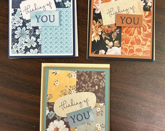 Könnte beinhalten: Drei handgefertigte Grußkarten mit floralen Mustern und dem Text "Thinking of You". Jede Karte hat ein einzigartiges Patchwork-Design mit verschiedenen Farben, darunter Blau, Orange, Gelb und Weiß. Die Karten sind rechteckig und haben einen weißen Rand.