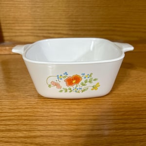 May include: White square casserole dish with a floral design featuring orange, pink, yellow, and blue flowers. The dish has two small handles and is set against a wooden background. A vintage kitchenware item.