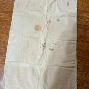 May include: Off-white, rectangular cloth with visible stains and wear. The fabric appears to be folded into three sections. The edges show signs of fraying. The cloth is lying on a wooden surface.