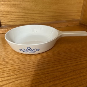May include: White ceramic skillet with a long handle. The skillet has a rounded bowl shape and is decorated with a blue floral pattern around the rim. The handle is a solid white color. The skillet is sitting on a wooden surface.