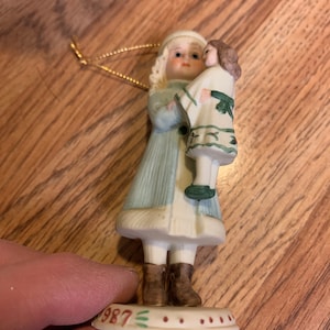 Könnte beinhalten: Eine Porzellanfigur eines Mädchens in einem blauen Mantel, das eine Puppe hält. Der Anhänger hat eine goldene Schnur zum Aufhängen und die Basis ist mit dem Jahr 1987 gekennzeichnet.