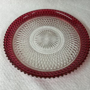 May include: A clear glass plate with a ruby red rim. The rim has a textured, diamond-like pattern. The center of the plate has a circular design with a starburst pattern. The plate is on a white surface.
