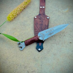 May include: A handcrafted dagger with a silver blade and a brown leather-wrapped handle. The blade features etched runes. It rests on a textured surface next to a brown leather sheath and a yellow plant. The sheath has a decorative design.
