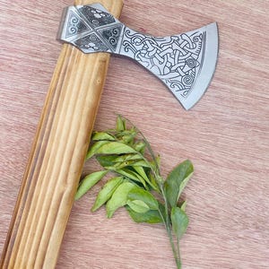 May include: A silver axe with a wooden handle. The axe head has a detailed design of a knotwork pattern. The handle is made of wood and has a smooth finish.