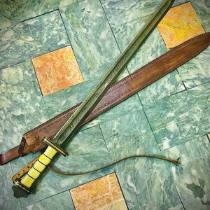 May include: A long, silver sword with a brown leather scabbard and a white handle. The sword is lying on a green and white marble floor.