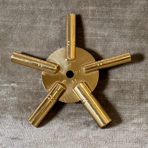 May include: A gold-colored watch key with a circular center and five cylindrical arms radiating outwards. The key has the numbers "5", "9", and "11" inscribed on the center. The key is made of brass or a similar metal.