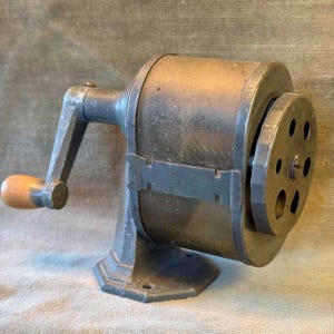 Può includere: Un temperamatite antico con un corpo in metallo grigio scuro e un manico in legno. Il temperamatite ha una forma cilindrica con una lama circolare e una manovella per il funzionamento manuale. La base è ottagonale.