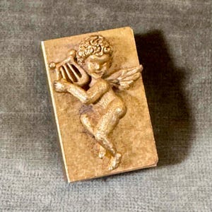 May include: A rectangular, gold-colored decorative plaque featuring a sculpted cherub playing a harp. The cherub has curly hair and small wings. The plaque is likely made of a composite material.