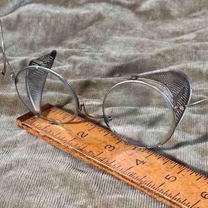 May include: Vintage steampunk-style safety glasses with clear round lenses and perforated metal side shields. The glasses are set on a wooden ruler, showcasing their size and design. The frame is made of silver-toned metal.