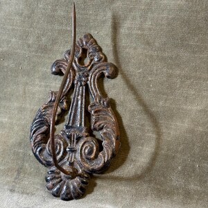 Pode incluir: Gancho de parede ornamentado, de estilo antigo, com acabamento em bronze. O gancho apresenta um design decorativo de volutas com um gancho curvo que se estende do centro. O metal tem uma aparência envelhecida, adicionando ao seu charme vintage.