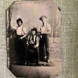 Può includere: Una fotografia vintage in bianco e nero di tre uomini in piedi davanti a un muro di mattoni. Gli uomini indossano cappelli e bretelle. L'uomo a sinistra tiene la mano sul fianco. L'uomo al centro è seduto su una sedia. L'uomo a destra tiene una bottiglia in mano.