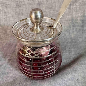 May include: A glass jar with a silver lid and handle, filled with a dark red preserve. The jar has a silver grid design around the outside. A silver spoon rests on the lid.