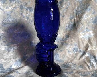 Può includere: Un calice di vetro blu cobalto dal design unico. Lo stelo presenta una mano scolpita che afferra la base della coppa. Il calice ha una base rotonda e un bordo svasato. Il vetro ha una finitura lucida.