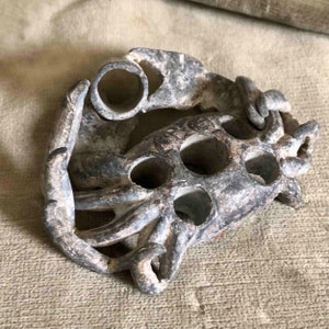 Op de afbeelding: Een verweerd, grijs metalen object met een reeks cirkelvormige gaten en een gebogen, organische vorm. Het object lijkt een decoratief stuk of een onderdeel van een grotere structuur te zijn. Het oppervlak vertoont tekenen van ouderdom en slijtage.