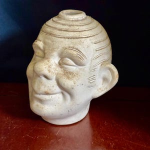 May include: A white ceramic head-shaped vase with a matte finish and a weathered, speckled texture. The sculpture features a stylized face with closed eyes, a prominent nose, and a small mouth, topped with a circular opening at the crown.