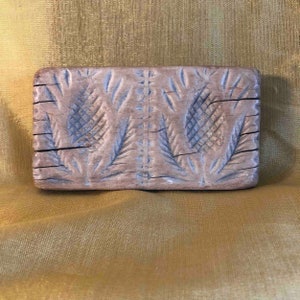Puede incluir: Un molde para galletas de madera vintage con un diseño tallado de dos piñas y hojas. El molde es de madera marrón claro y tiene forma rectangular.