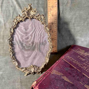 May include: An ornate oval-shaped brass picture frame with a delicate floral design. The frame has a mesh backing and is approximately 5 inches tall.
