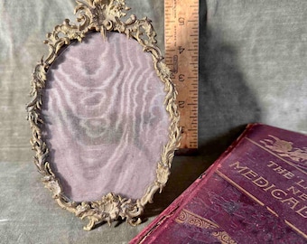 May include: An ornate oval-shaped brass picture frame with a delicate floral design. The frame has a mesh backing and is approximately 5 inches tall.