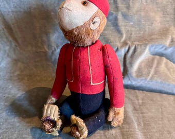 Puede incluir: Un muñeco de mono de peluche vintage con una chaqueta y gorra rojas, con pantalones azul oscuro. El mono tiene una cara blanca, pelaje marrón y manos y pies color canela. El muñeco está sentado con las piernas dobladas.