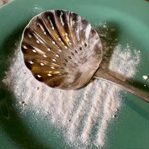 May include: A vintage silver-toned strainer with a shell-shaped bowl and a long handle rests on a green surface dusted with white powder. The strainer has decorative cut-out holes.
