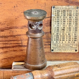 Op de afbeelding: Een cilindrisch messing gereedschapsonderdeel met een uitlopende voet en een bovengedeelte met twee ronde openingen aan de zijkant. Het object staat naast een metalen plaat met een tabel van decimale equivalenten.