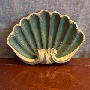 May include: A teal-coloured, shell-shaped wall hanging. The shell has a ribbed texture and a cream-coloured trim. The piece is made of a solid material and is likely used for decorative purposes.